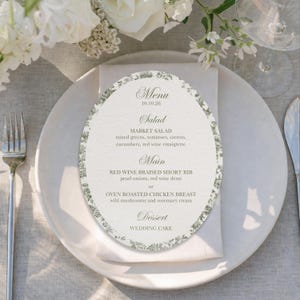 May include: A wedding menu card on a white plate, featuring a floral border and a list of dishes. The menu includes salad, main courses, and dessert options. The date on the menu is 10.10.26. Silverware and flowers are in the background.