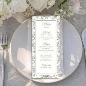 May include: A formal menu card with a floral border sits on a white plate with a napkin. The menu lists a salad, main course options of short rib or chicken breast, and wedding cake for dessert. The date 10.10.26 is at the top.