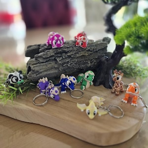 Könnte beinhalten: Eine Sammlung farbenfroher Tier-Schlüsselanhänger, darunter ein Panda, Dinosaurier, eine Giraffe und eine Schildkröte. Die Schlüsselanhänger bestehen aus einem harten Material und sind an silbernen Schlüsselringen befestigt. Sie sind auf einem Holzbrett angeordnet.