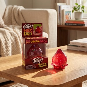 May include: A Dr. Pepper Nee Doh Dream Drop toy, a red, teardrop-shaped stress ball with glitter inside, sits on a wooden table. The toy is next to its box, which features the Dr. Pepper logo and the words "Suptid Squish!"