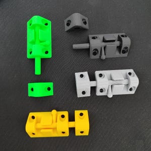 Könnte beinhalten: Eine Auswahl an 3D-gedruckten Türriegeln in Grün, Grau und Gelb. Die Riegel verfügen über einen Schieberiegelmechanismus und Befestigungslöcher. Eine Eckhalterung ist ebenfalls vorhanden. Maße in Zentimetern.