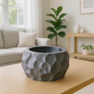 May include: A gray, textured planter with a unique, organic design. The planter is round with a wide opening and sits on a light-colored wooden table. The interior of the planter is a darker gray.