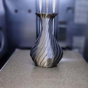 Pode incluir: Um vaso preto e prateado com um design em espiral. O vaso tem uma base larga que se afunila para um pescoço estreito e uma borda alargada. O vaso está sobre uma superfície cinzenta, com uma impressora 3D no fundo.