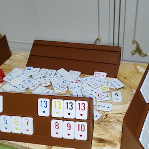 May include: A board game setup with brown wooden holders and white tiles featuring numbers and symbols. A red die sits near the tiles. The game is set on a light-colored wooden surface.