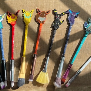 May include: A collection of makeup brushes with character-themed toppers. The brushes feature various colors and designs, including green, blue, red, yellow, orange, black, and white. The brushes are arranged on a wooden surface.