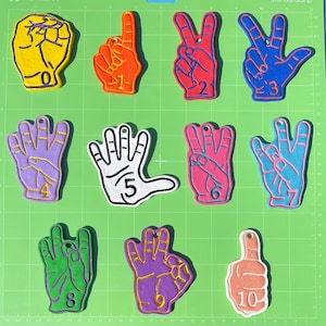 May include: A collection of colorful hand-shaped ornaments, each displaying a number from zero to ten. The ornaments are in various colors, including yellow, orange, red, blue, purple, white, pink, and green, with gold or white outlines. They are arranged on a green cutting mat.
