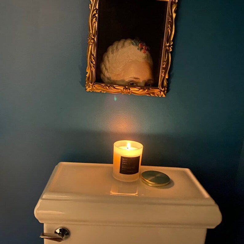 May include: A lit candle in a clear glass jar with a black label sits on a white surface. A small, round, green lid is next to the candle. Above, a framed portrait of a person with a powdered wig hangs on a teal wall.