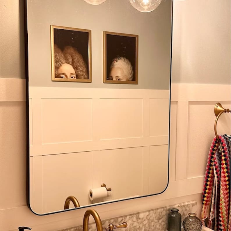 May include: Two framed portraits of figures with elaborate hairstyles and partially visible faces hang above a white paneled wall. The portraits are in gold frames. A large mirror reflects the bathroom fixtures.