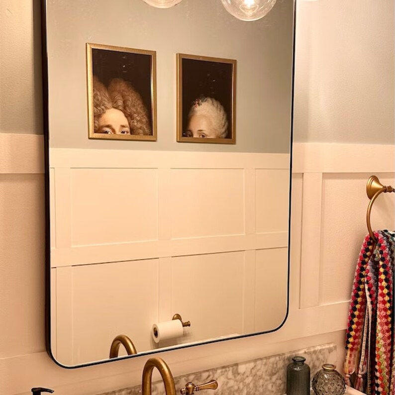 Pu&ograve; includere: Un bagno con uno specchio con cornice nera, boiserie bianca e accessori dorati. Due ritratti incorniciati di persone con parrucche sono appesi al muro sopra lo specchio.