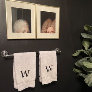 Pu&ograve; includere: Due asciugamani bianchi con la lettera "W" ricamata su di essi sono appesi a un portasciugamani cromato in un bagno nero. Due ritratti incorniciati di persone con parrucche sono appesi al muro dietro gli asciugamani.