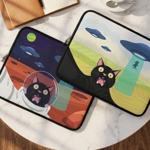 May include: Two rectangular laptop sleeves featuring cartoon illustrations of black cats and UFOs. One sleeve shows a cat in a space helmet on a red planet, while the other depicts a cat being abducted by a flying saucer in a green field.