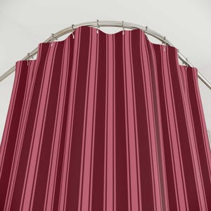May include: A shower curtain featuring a vertical striped pattern in alternating shades of deep burgundy and muted rose pink. The fabric hangs from a curved silver metal rod with visible circular rings, creating soft folds across the entire surface.