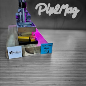 Op de afbeelding: Een metalen en houten pers met de tekst "Pix2Mag" op een wit label en "Your Logo Here" op een blauw label. De pers heeft een zwart, zilver en goud kleurenschema, met een roze accent. De achtergrond bevat de tekst "Ply2Mag".
