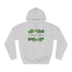 Könnte beinhalten: Ein hellgrauer Kapuzenpullover mit einer Vordertasche und einer Kapuze mit Kordelzug. Der Pullover zeigt den Text "Plant Mom" in eleganter Schrift, umrahmt von grünen tropischen Blättern im Aquarellstil.