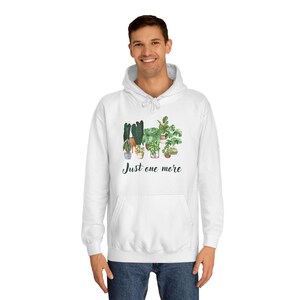 Könnte beinhalten: Weißer Kapuzenpullover mit einer Aquarellillustration verschiedener Topfpflanzen. Der Schriftzug "Just one more" steht unter der Pflanzengrafik. Der Pullover hat eine Kapuze mit Kordelzug und eine Vordertasche.
