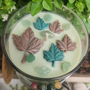 Puede incluir: Una vela de cristal transparente con cera verde claro. La vela está decorada con formas de hojas de arce marrones y verde azulado y piedras verdes. La vela está frente a follaje verde.