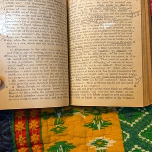 May include: An open book with aged, yellowed pages, featuring handwritten notes and printed text. The book is titled "ESSAYS" and discusses Shakespeare. The background includes a colorful, patterned quilt.