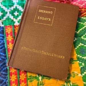 May include: A brown hardcover book titled "Emerson's Essays" from Macmillan's Pocket Classics. The book is set against a colorful patchwork quilt background with green, yellow, pink, and blue patterns. The book's cover has gold lettering.