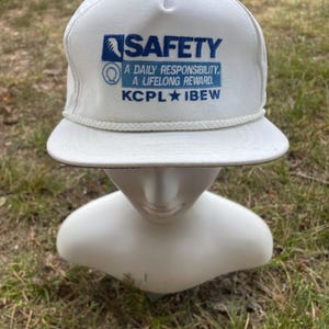May include: White baseball cap with the word "SAFETY" in blue block letters. The cap also features the text "A DAILY RESPONSIBILITY, A LIFELONG REWARD. KCPL IBEW" in blue. The cap has a white rope detail and a curved brim.