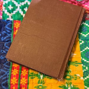 May include: A brown hardcover book with a textured cover. The book is slightly angled, revealing the spine and edges. The background is a colorful patchwork quilt with various patterns and colors.