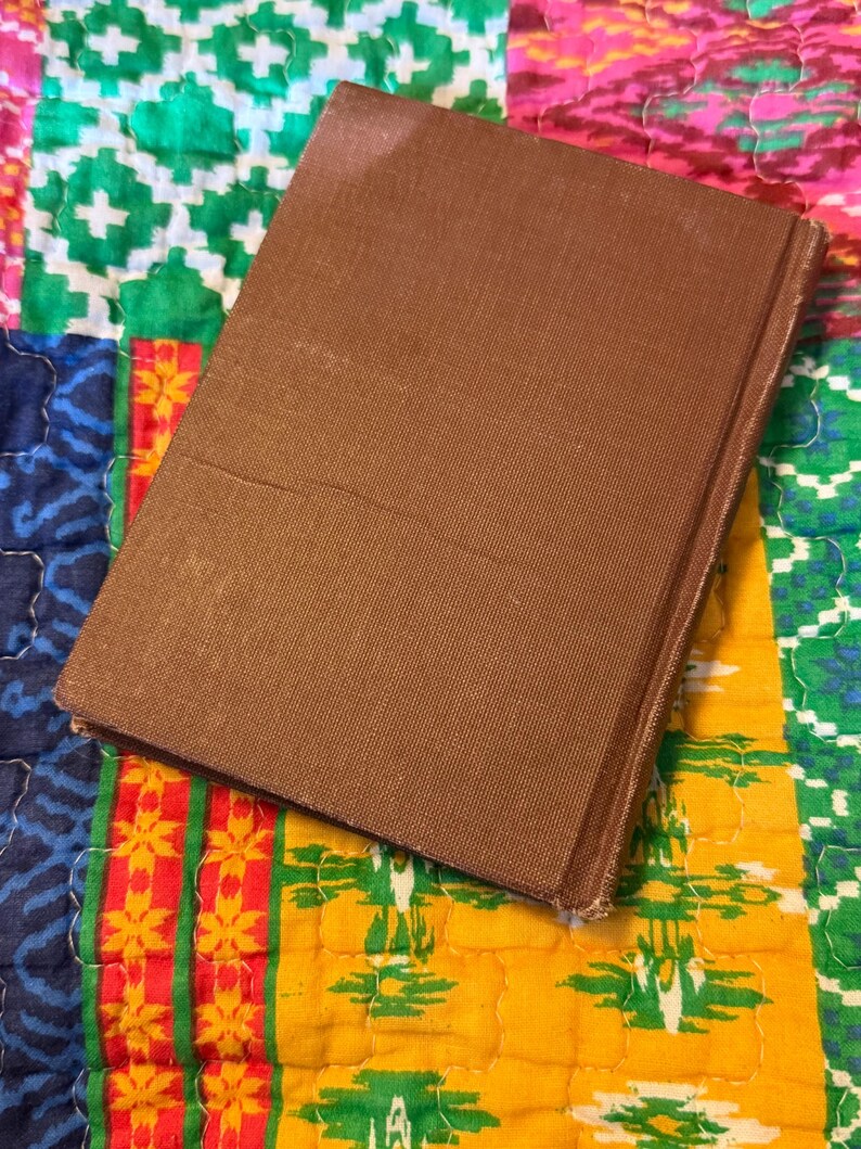 May include: A brown hardcover book with a textured cover. The book is slightly angled, revealing the spine and edges. The background is a colorful patchwork quilt with various patterns and colors.