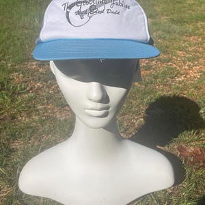 May include: White and blue trucker hat with the text "The Goodtime Jubilee" and a guitar illustration. The hat has a white front panel, a blue brim, and a mesh back. The text "Mrs. Hazel Dudd" is also printed on the hat.