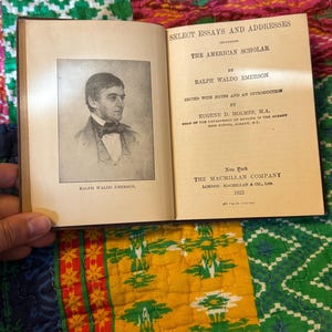 May include: An open book with the title "Select Essays and Addresses" and "The American Scholar" by Ralph Waldo Emerson. A portrait of Emerson is on the left page. Published by The Macmillan Company in 1922.
