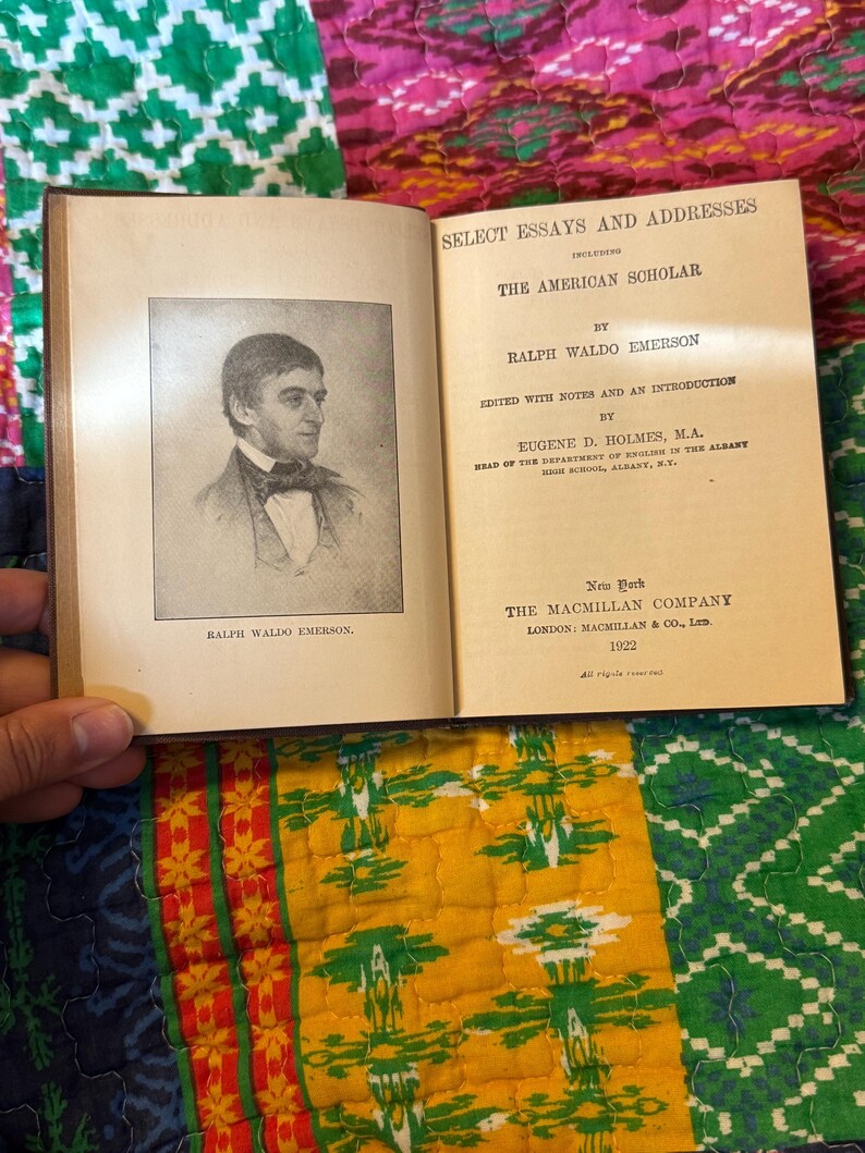 May include: An open book with the title "Select Essays and Addresses" and "The American Scholar" by Ralph Waldo Emerson. A portrait of Emerson is on the left page. Published by The Macmillan Company in 1922.