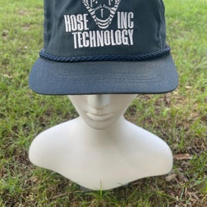 May include: Navy blue cap with the text "HOSE INC TECHNOLOGY" in white. The cap has a curved brim and a rope detail along the base. The cap is displayed on a white mannequin head.