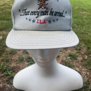 Puede incluir: Gorra de camionero gris con visera curva. La gorra presenta texto y gráficos bordados, incluidas las letras "NRA" e "ILA", junto con la cita, "That every man be armed."