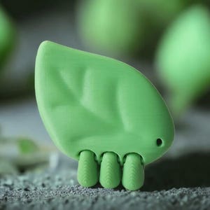 Puede incluir: Objeto decorativo en forma de hoja verde claro, con tres pequeñas patas texturizadas. El objeto tiene una superficie lisa con detalles sutiles de venas y un pequeño punto oscuro que sugiere un ojo. El fondo presenta formas verdes borrosas.