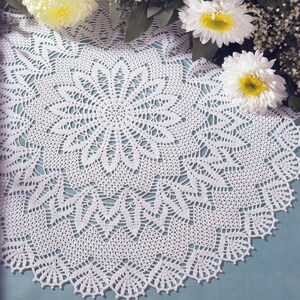 Puede incluir: Un tapete blanco circular de ganchillo con un detallado patrón floral. Flores blancas y amarillas con hojas verdes rodean el tapete, creando una estética delicada y artesanal. El intrincado diseño es visible en toda la pieza.
