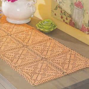 Könnte beinhalten: Ein gehäkelter Tischläufer in Pfirsichfarbe mit einem zarten Blumenmuster. Der Läufer liegt auf einem Holztisch mit einer weißen Vase mit rosa Blumen und einem grünen Dekorationsobjekt.