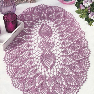 May include: A delicate, oval-shaped crochet doily in a light purple color. The doily features a intricate design with a central motif and radiating patterns.