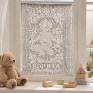 May include: A white rectangular crochet window panel featuring a central motif of a cherub surrounded by floral patterns and the name SOPHIA crocheted in block letters at the bottom. The intricate lace design is displayed against a window pane.