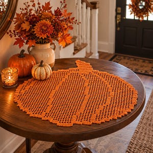 Op de afbeelding: Een oranje gehaakt kleedje in de vorm van een pompoen met een rasterpatroon. Het kleedje ligt op een ronde houten tafel naast een kleine brandende kaars, een grote oranje pompoen, een kleine witte pompoen en een keramieken vaas met herfstbladeren.