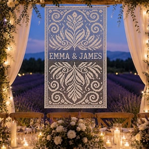 May include: A rectangular white lace hanging featuring an intricate botanical pattern of leaves and swirls. The text EMMA & JAMES is centered horizontally in a bold, sans-serif font within the lace design, which is suspended from a wooden beam.