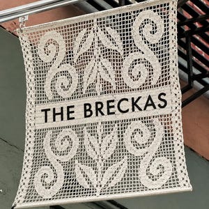 Op de afbeelding: Een rechthoekige wandhanger van gebroken wit haakwerk met een opengewerkt rasterpatroon en massieve blad- en spiraalmotieven. De tekst THE BRECKAS staat in zwarte hoofdletters op een horizontale band in het midden van het object.