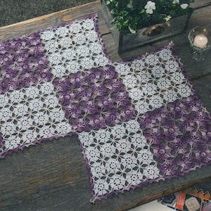 May include: Two handmade crochet placemats. Each placemat features a white and purple floral pattern. The placemats are made of a soft, textured yarn and have a scalloped edge. The placemats are displayed on a wooden surface.