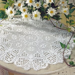 Peut inclure: Nappe ronde blanche en crochet avec un motif ajouré détaillé. La nappe est posée sur une surface claire et est entourée de marguerites blanches et de feuillage vert, créant une esthétique délicate et vintage.