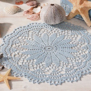May include: A light blue crocheted doily with a delicate floral pattern. The doily is laid out on a white wooden surface with seashells and a starfish.