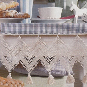 Può includere: Un runner da tavolo in crochet bianco con un motivo a quadri blu e bianco. Il runner ha un bordo smerlato con frange e un motivo a rombi.