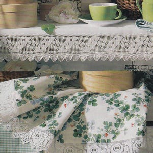May include: A still life arrangement featuring a white tablecloth with a delicate lace trim. The table is adorned with a green teacup and saucer, a floral print fabric, and decorative wooden boxes. A basket and a floral artwork are in the background.