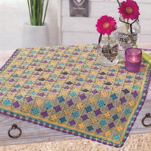 May include: A handmade, square crochet blanket in a neutral colour palette with a repeating floral pattern. The blanket features a border of teal, yellow, and purple. The blanket is displayed on a wooden surface.