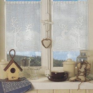 May include: A window with white crocheted curtains showing deer silhouettes. A small birdhouse, a cup and saucer, and a jar with heart-shaped decorations are on the windowsill. A blue and white patterned cloth is also present.