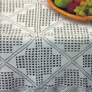 May include: A white, crocheted tablecloth with a geometric pattern and scalloped edge. The intricate design features squares and diamond shapes. A woven basket filled with green and red apples and green grapes sits on the corner of the tablecloth.