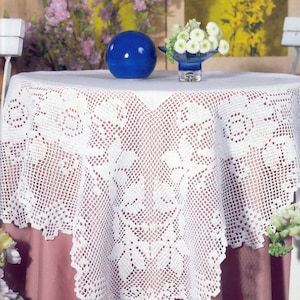 May include: A white, crocheted tablecloth with a detailed floral pattern covers a round table. A blue ceramic sphere and a small vase of white flowers sit on the table. The tablecloth's edge has a scalloped design.