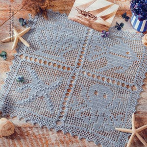 Peut inclure: Nappe ajourée en crochet bleu clair, avec un motif détaillé. Autour, des étoiles de mer, des coquillages et une carte postale d'un voilier. L'ensemble est posé sur une surface en bois, évoquant un thème marin.