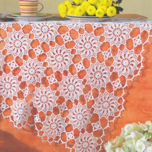 May include: A white, crocheted lace tablecloth with a floral pattern covers an orange surface. A yellow and red striped mug sits on a saucer. A plate of yellow flowers is on the table, and white flowers are in the foreground.