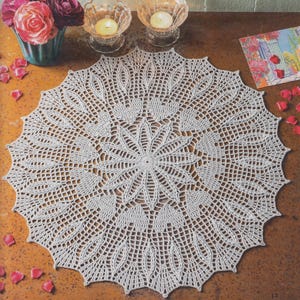 May include: A white, circular, crocheted doily with a detailed, openwork design. The doily features a central starburst pattern surrounded by leaf-like motifs. The doily is approximately 20 inches in diameter. The image also includes flowers, candles, and a card.